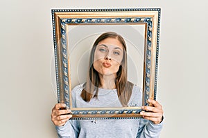 Beautiful caucasian woman holding empty frame looking at the camera blowing a kiss being lovely and sexy