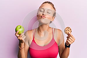 Beautiful caucasian woman holding green apple and cookie looking at the camera blowing a kiss being lovely and sexy