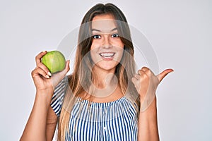 Beautiful caucasian woman holding green apple pointing thumb up to the side smiling happy with open mouth