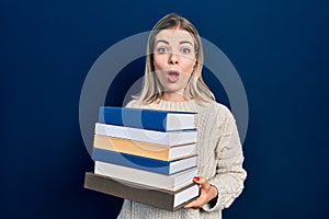 Beautiful caucasian woman holding a pile of books afraid and shocked with surprise and amazed expression, fear and excited face