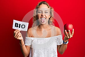 Beautiful caucasian woman holding red apple and bio word sticking tongue out happy with funny expression
