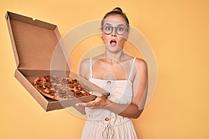 Beautiful caucasian woman holding tasty pepperoni pizza afraid and shocked with surprise and amazed expression, fear and excited