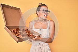 Beautiful caucasian woman holding tasty pepperoni pizza angry and mad screaming frustrated and furious, shouting with anger