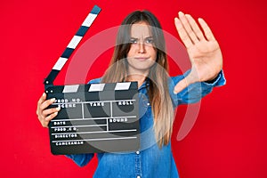 Beautiful caucasian woman holding video film clapboard with open hand doing stop sign with serious and confident expression,