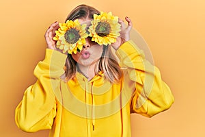 Beautiful caucasian woman holding yellow sunflowers over eyes making fish face with mouth and squinting eyes, crazy and comical