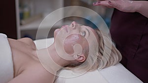 Beautiful Caucasian woman lying in spa salon as cosmetician applying moisturizing facial cream in slow motion after