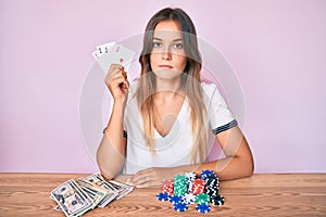 Beautiful caucasian woman playing poker holding cards thinking attitude and sober expression looking self confident