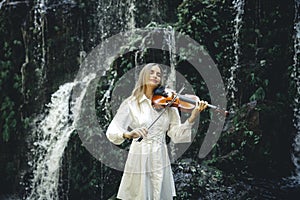 Beautiful Caucasian woman playing violin near waterfall. Music and art concept. Charming female wearing white dress in nature.