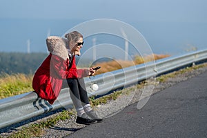 Beautiful Caucasian woman with red coat sit on barrier of roadside and use mobile phone on mountain near windmill or wind turbine