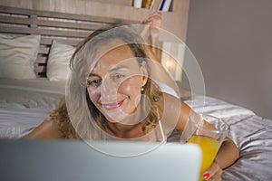 Beautiful Caucasian woman 30s lying on bed at night in home bedroom using internet at laptop computer drinking healthy orange juic