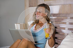 Beautiful Caucasian woman 30s sitting on bed at night in home bedroom using internet at laptop computer drinking healthy orange ju
