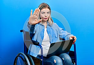 Beautiful caucasian woman sitting on wheelchair using laptop with open hand doing stop sign with serious and confident expression,