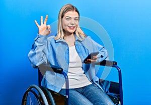 Beautiful caucasian woman sitting on wheelchair using smartphone doing ok sign with fingers, smiling friendly gesturing excellent