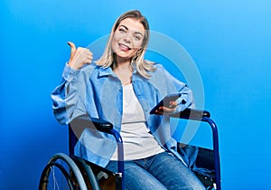 Beautiful caucasian woman sitting on wheelchair using smartphone pointing thumb up to the side smiling happy with open mouth