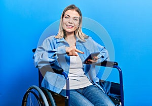 Beautiful caucasian woman sitting on wheelchair using smartphone smiling happy pointing with hand and finger