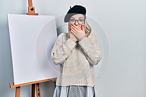 Beautiful caucasian woman standing by painter easel stand shocked covering mouth with hands for mistake