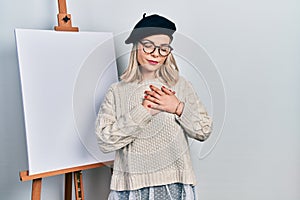 Beautiful caucasian woman standing by painter easel stand smiling with hands on chest with closed eyes and grateful gesture on