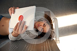 Beautiful caucasian woman in trendy casual clothes holding a tablet. Confident girl lying down on iron seats in a waiting room wat