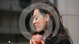 Beautiful Caucasian woman walking around grey building, smiling, looking at her phone, eating tasty big apple.