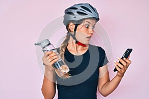 Beautiful caucasian woman wearing bike helmet looking at smartphone afraid and shocked with surprise and amazed expression, fear