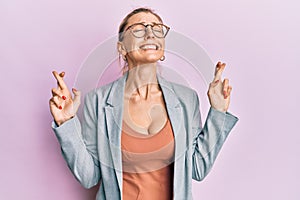 Beautiful caucasian woman wearing business jacket and glasses gesturing finger crossed smiling with hope and eyes closed