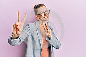 Beautiful caucasian woman wearing business jacket and glasses smiling looking to the camera showing fingers doing victory sign