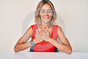 Beautiful caucasian woman wearing casual clothes sitting on the table smiling with hands on chest with closed eyes and grateful