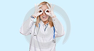 Beautiful caucasian woman wearing doctor uniform and stethoscope doing ok gesture like binoculars sticking tongue out, eyes