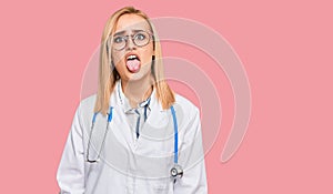 Beautiful caucasian woman wearing doctor uniform and stethoscope sticking tongue out happy with funny expression