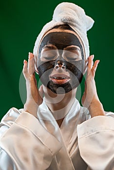Beautiful caucasian woman in white towel applying black clay mask on her face isolated on green