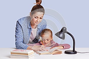 Beautiful Caucasian woman helping her doughter to do school homework, mother and child surronded by books, little girl sitting
