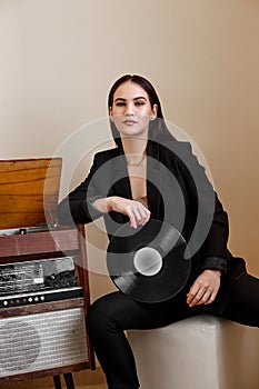 A beautiful Caucasian young woman in a black pantsuit and black sandals poses next to a vintage record player with a gramophone