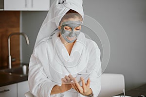 Beautiful caucasian young woman with facial mask look at screen of mobile phone