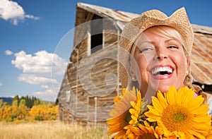 Beautiful Caucasian Young Woman Wearing Cowboy Hat Holding Sunflower In Front of Rustic Barn In The Country