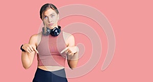 Beautiful caucasian young woman wearing gym clothes and using headphones pointing down looking sad and upset, indicating direction