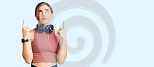 Beautiful caucasian young woman wearing gym clothes and using headphones pointing up looking sad and upset, indicating direction