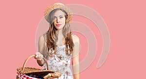 Beautiful caucasian young woman wearing summer hat and holding picnic wicker basket with bread thinking attitude and sober