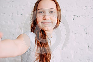 Beautiful charming redhead smiling teenager girl taking selfie on frontal camera isolated on white brick wall