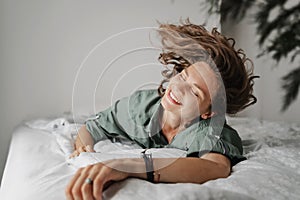 Beautiful charming young curly girl woman in a green shirt lies on the bed, shot of a happy smiling face and flying hair