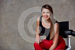 A beautiful cheeky young girl sits in a chair and smiles in a chamber against a gray wall. The concept of a successful