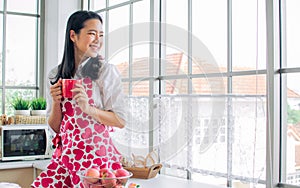 Beautiful cheerful Asian woman wearing apron, smiling with happiness, drinking and holding cup of coffee or tea, standing in