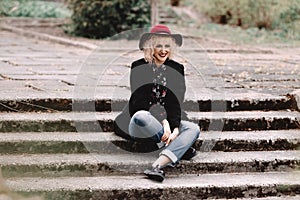 Beautiful cheerful blonde girl with short curly hair in coat and hat sitting on stairs on the street