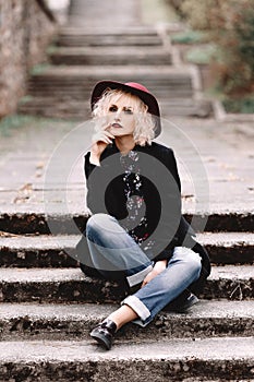 Beautiful cheerful blonde girl with short curly hair in coat and hat sitting on stairs on the street