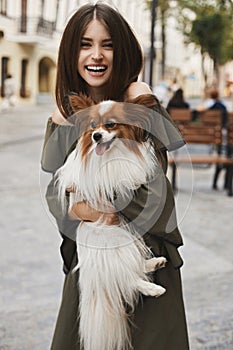 Beautiful and cheerful brunette model girl with shiny smile, in short dress with a small cute papillon dog on her hands posing out