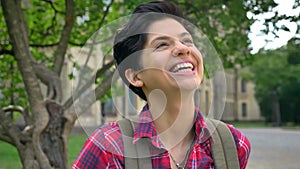 Beautiful cheerful college student with black short hair laughing and smiling at camera, standing in park near
