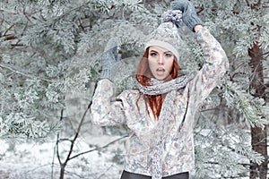 Beautiful cheerful cute red-haired girl in a cap and scarf plays with snow in the beautiful forest fairy