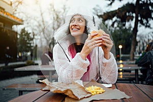 Beautiful and cheerful girl eating a juicy hamburger and french fries on the street