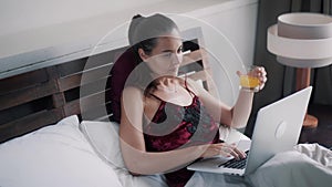 Beautiful cheerful girl works on laptop drinking orange juice in bed