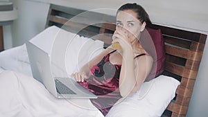 Beautiful cheerful girl works on laptop drinking orange juice in bed