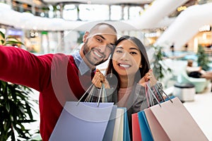 Beautiful cheerful interracial couple holding paper bags, taking selfie at mall, going shopping together